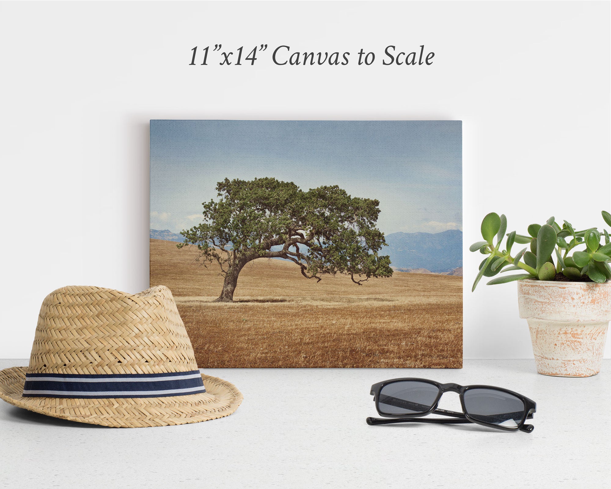 Canvas print of a solitary tree in a field against a mountain backdrop, placed on a surface. A straw hat, sunglasses, and potted plant are arranged in front of the canvas. Text above the canvas reads "11x14 Rustic Canvas Print (Choose from 10+ Designs)" by Offley Green. Perfect for those who appreciate rustic wall art.