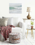 A cozy living room includes a white sofa with patterned cushions and a red geometric throw, complemented by a round pouf. A side table holds a lamp, pitcher, and plant. The wall is adorned with Offley Green's 16x20 Coastal Canvas Prints, adding to the seaside vibe with various designs.