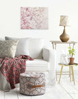 A cozy living room featuring a white sofa with patterned pillows and a red geometric throw blanket, complemented by a round patterned pouf. Nearby, there's a side table with a lamp, green plant, and glass pitcher. An Offley Green 16X20 Botanical Canvas Print adorns the white wall.