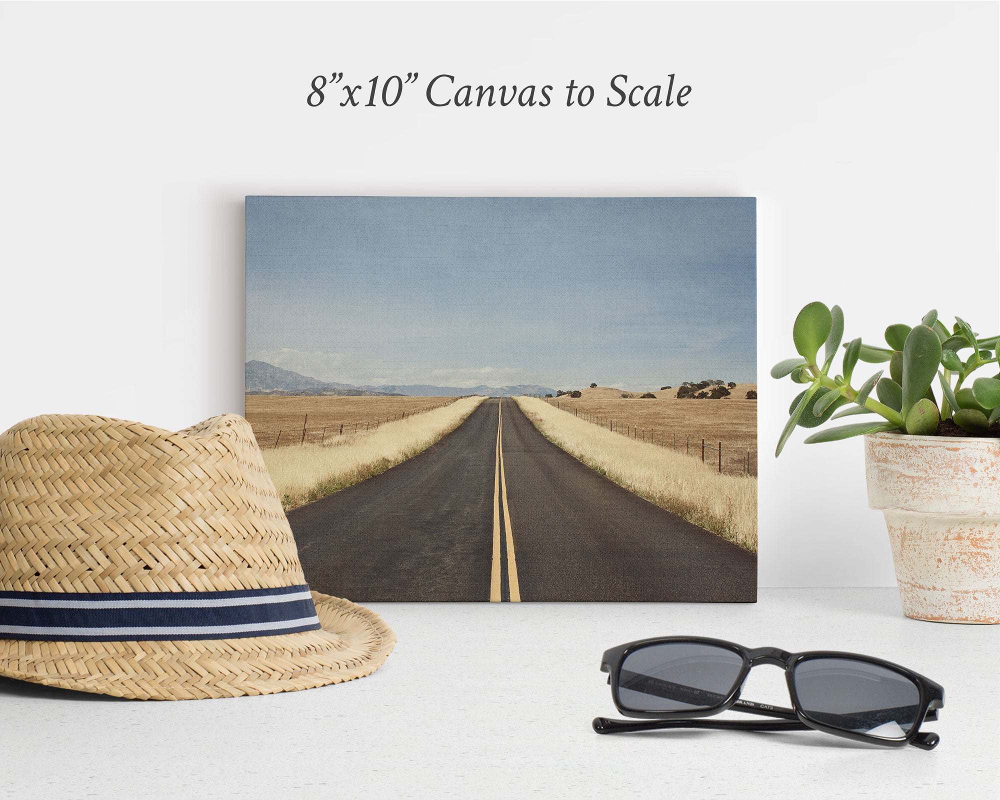 The Offley Green 8x10 Rustic Canvas Print features a straight road in a rural landscape. Nearby, charm is added with a straw hat, black sunglasses, and potted plant. Text above states "8x10 Canvas to Scale." Choose from 10+ designs.