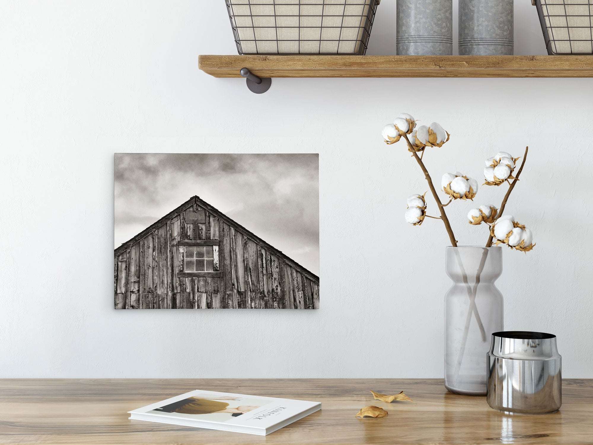 A rustic room has a wooden table with a metal pot of cotton branches. A shelf above holds wire baskets and candles. An Offley Green 8x10 Rustic Canvas Print, featuring one of over 10 designs like black-and-white weathered barns, adds charm alongside a book and petals.