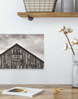 A rustic room has a wooden table with a metal pot of cotton branches. A shelf above holds wire baskets and candles. An Offley Green 8x10 Rustic Canvas Print, featuring one of over 10 designs like black-and-white weathered barns, adds charm alongside a book and petals.