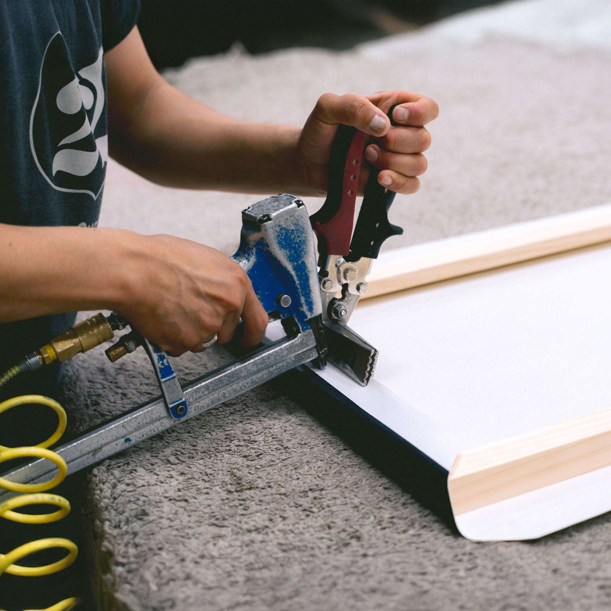 Using an Offley Green staple gun with a blue handle and connected by a yellow air hose, the person expertly secures a botanical print canvas from the "Set of 4 Framed Canvas 24x30" collection to a wooden frame on a soft surface.