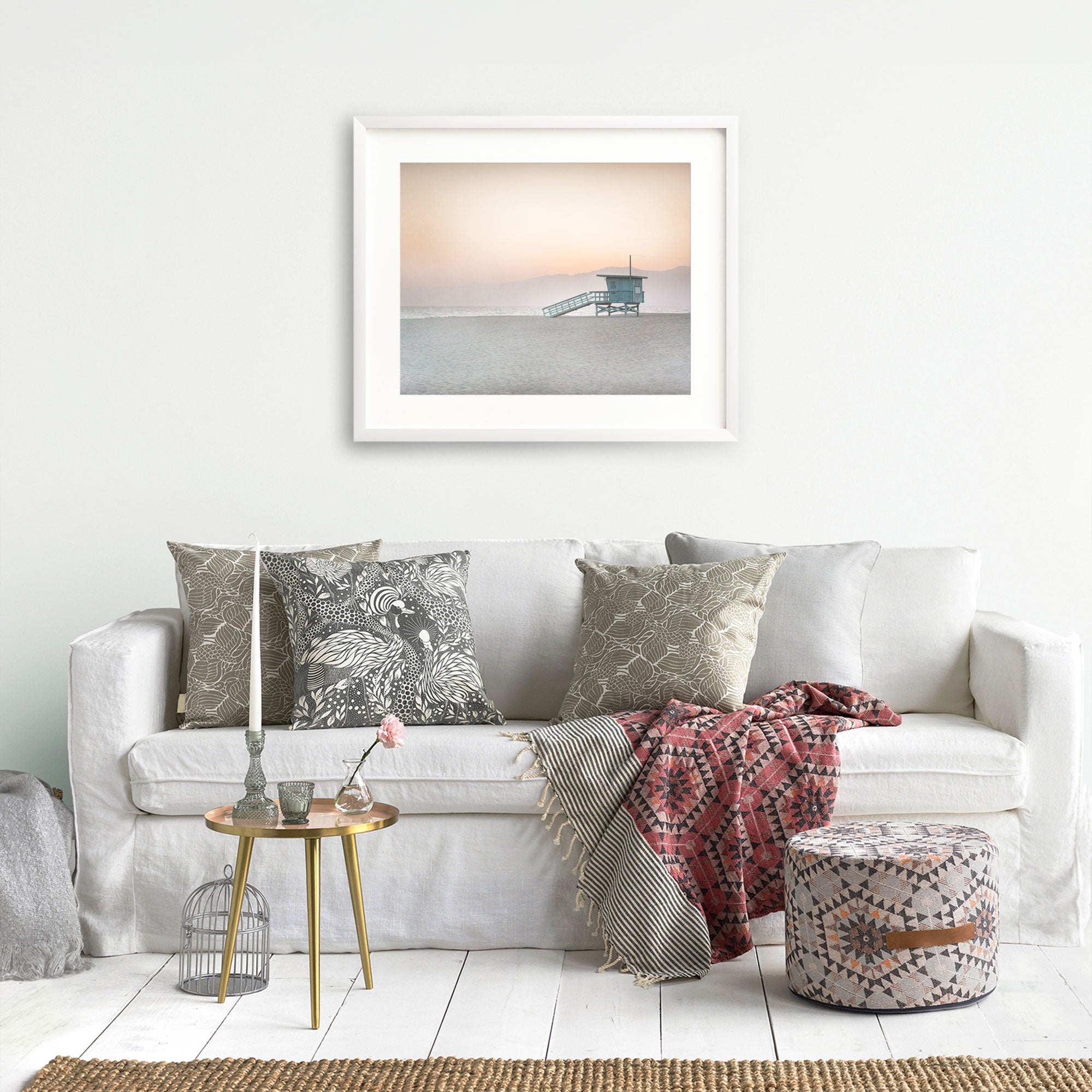 A cozy living room scene with a white sofa adorned with decorative pillows, a red patterned throw blanket, and a small table with vases and books. A tranquil Pink Coastal Print, 'Lifeguard Tower' photograph of Venice hangs on the wall from Offley Green.