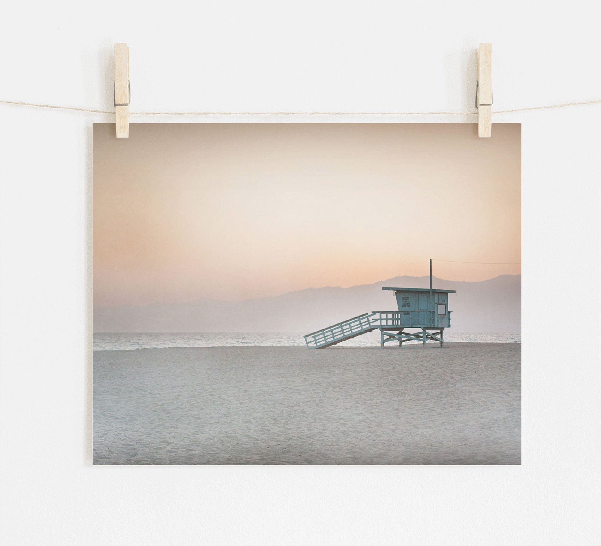 A serene beach scene at dusk printed on archival photographic paper, hung by clips on a string against a white wall. It features the Offley Green Pink Coastal Print, 'Lifeguard Tower' on an empty sandy beach near Venice and Santa Monica.