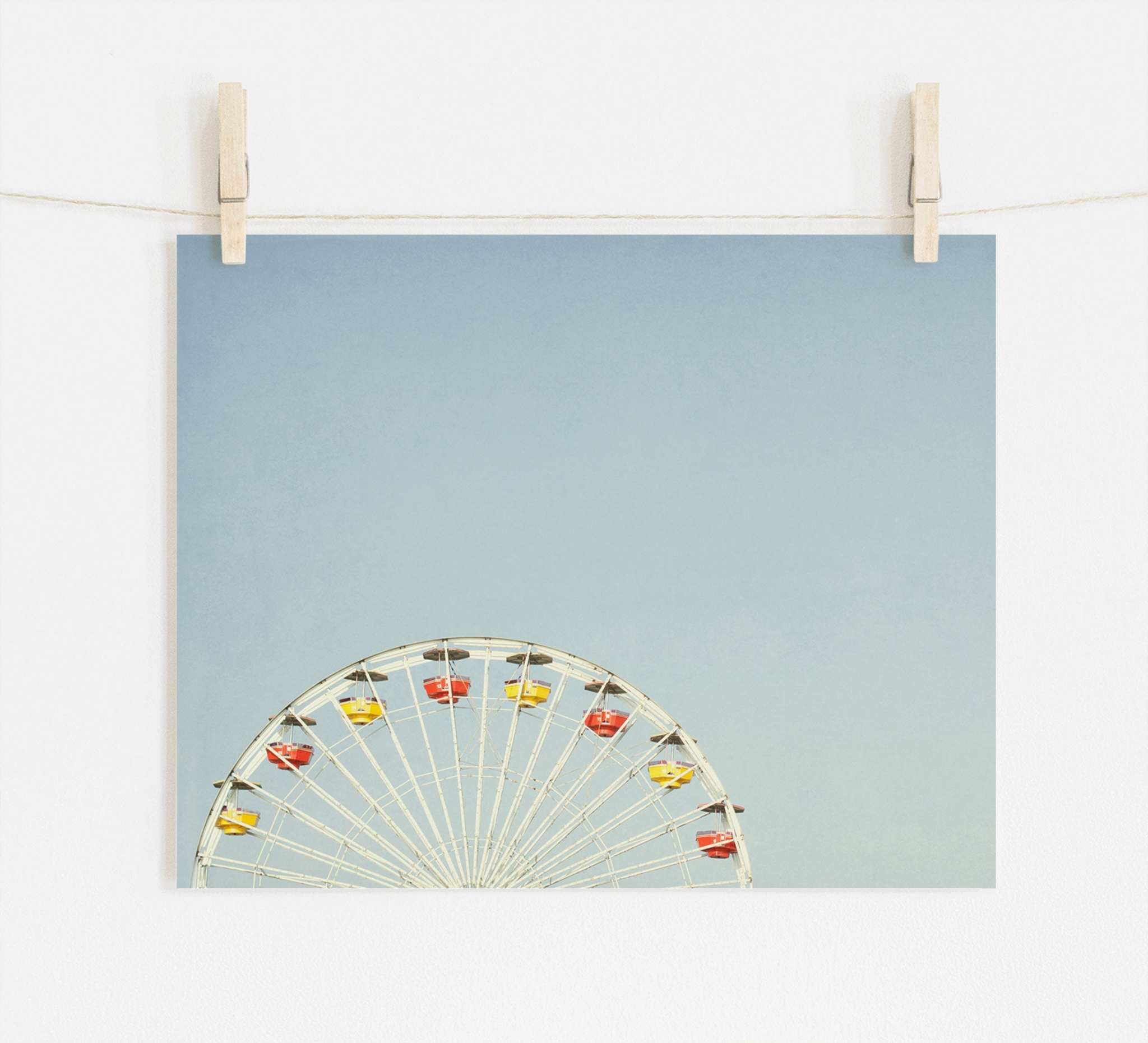 A photograph pinned by wooden clothespins on a string, depicting the top half of a ferris wheel with colorful cabins at Santa Monica Pier, set against a plain light blue background - Blue Minimalist Wall Decor, 'Ferris Blue' by Offley Green.