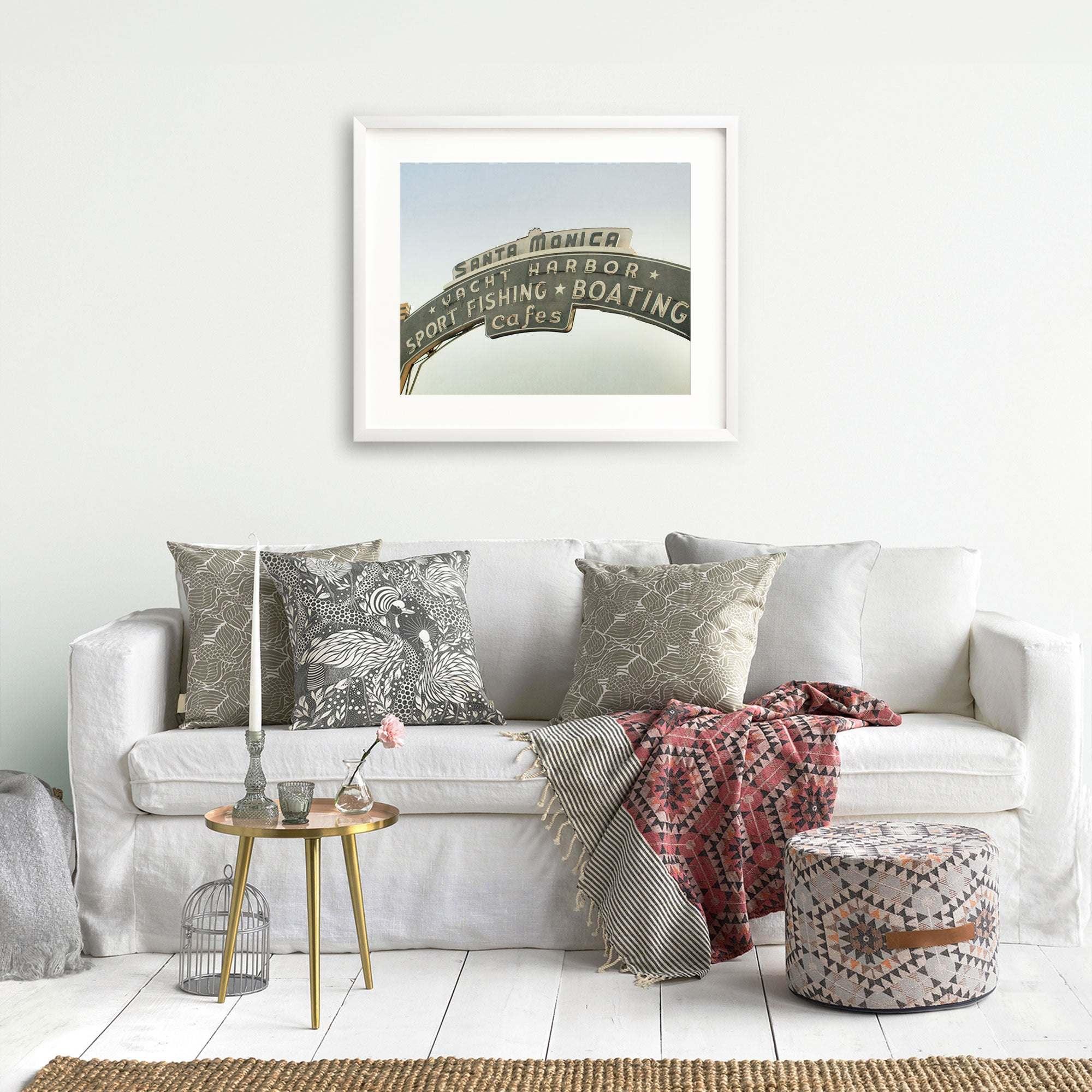 A cozy living room with a white sofa adorned with patterned cushions, an Offley Green Los Angeles California Print, 'Santa Monica Pier Blues' above it, a small round table with books and cups, and a colorful woven throw blanket.
