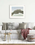 A cozy living room with a white sofa adorned with patterned cushions, an Offley Green Los Angeles California Print, 'Santa Monica Pier Blues' above it, a small round table with books and cups, and a colorful woven throw blanket.