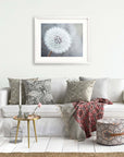 A cozy living room scene featuring a white sofa with decorative pillows, an unframed Neutral Grey Floral Print of a dandelion on the wall, a small round table with books, a birdcage, and 'Dandelion King' by Offley Green.