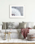 A cozy, inviting living room nook features a white sofa covered with decorative gray and beige pillows, a patterned red blanket, a small round table with books, flowers, and an Offley Green Grey Botanical Print, 'Dandelion Queen'.