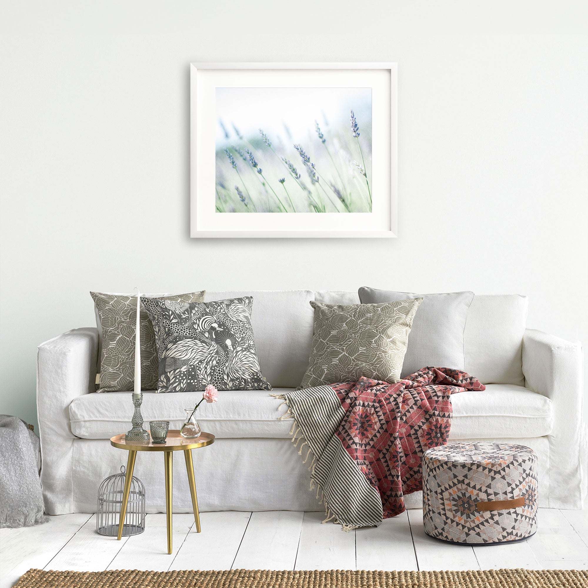 A cozy living room scene with a white sofa adorned with patterned cushions, a red and gray throw blanket, and a small gold side table. A framed picture of the Santa Ynez Valley hangs above the Offley Green Rustic Farmhouse Floral Wall Art, &#39;Buds of Lavender&#39;.