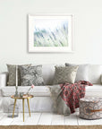 A cozy living room scene with a white sofa adorned with patterned cushions, a red and gray throw blanket, and a small gold side table. A framed picture of the Santa Ynez Valley hangs above the Offley Green Rustic Farmhouse Floral Wall Art, 'Buds of Lavender'.