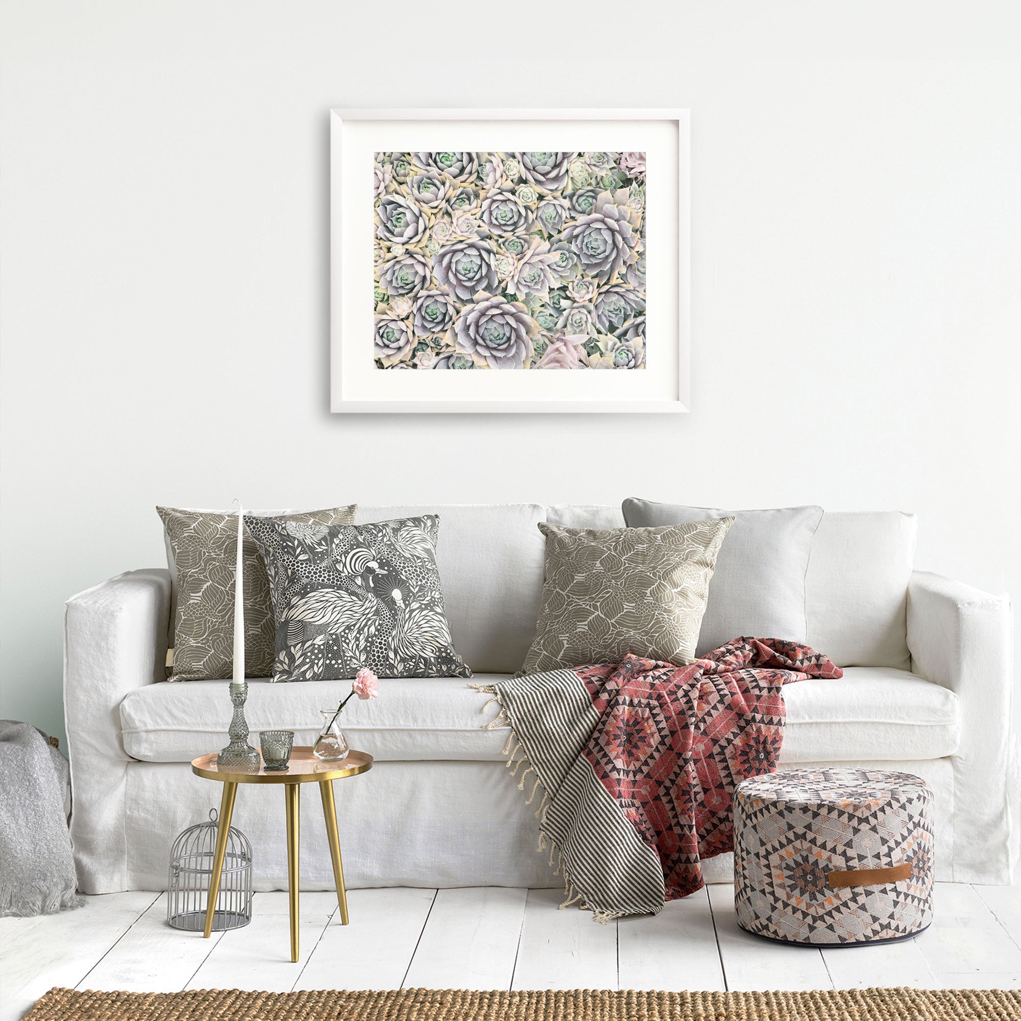A cozy living room with a white sofa adorned with various patterned cushions in neutral tones and a red geometric throw blanket. A small round wooden side table with a candle, flowers, and a metal lantern sits beside the sofa. An Offley Green framed Pastel Green Botanical Print 'Succulents Forever' on archival photographic paper hangs above.