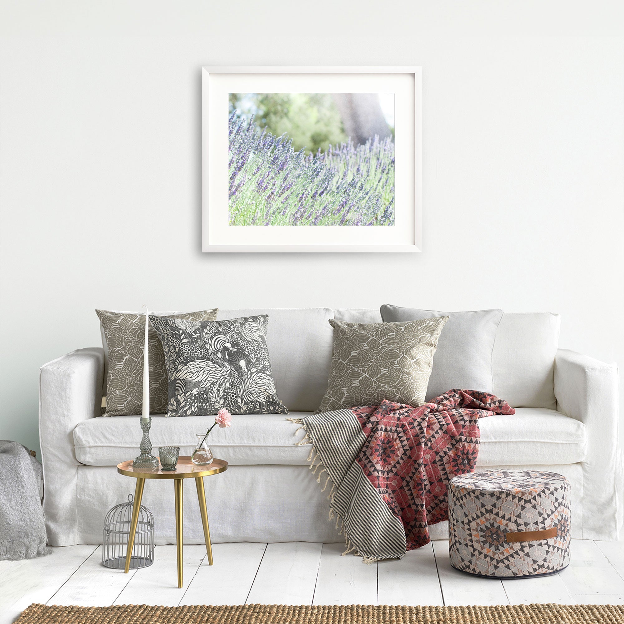 A cozy living room with a white sofa adorned with various patterned cushions and a pink and gray throw blanket. A round wooden side table with a vase and a candle sits beside the sofa. Unframed Rustic Floral Prints, 'Fields of Lavender,' by Offley Green hang above the sofa.