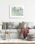 A cozy living room with a white sofa adorned with various patterned cushions and a pink and gray throw blanket. A round wooden side table with a vase and a candle sits beside the sofa. Unframed Rustic Floral Prints, 'Fields of Lavender,' by Offley Green hang above the sofa.