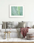 A cozy living room scene with a white couch adorned with patterned pillows, a red throw blanket, a small round table with a book and an Aloe Vera plant, a framed abstract art piece featuring the Mint Green Botanical Print 'Aloe Vera Spikes' from Offley Green.