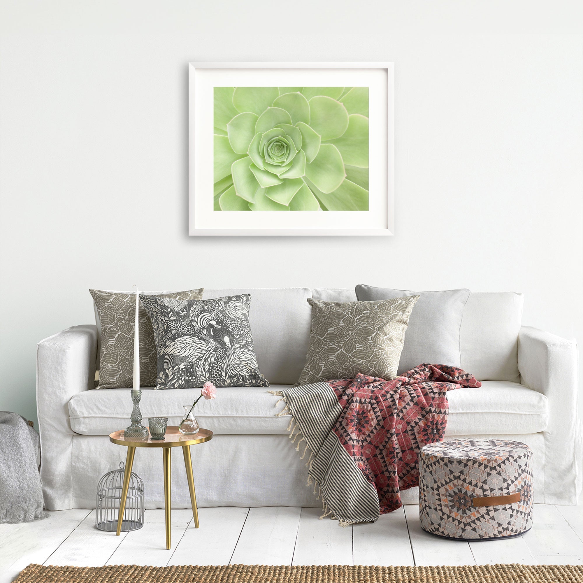 A cozy living room with a white sofa adorned with patterned cushions in various shades of grey and beige. A decorative throw blanket is draped over the sofa. A small round wooden table holds a pink flower in a glass vase. An ottoman and Offley Green's Sage Green Print, 'Succulent Heart' adorn the space.