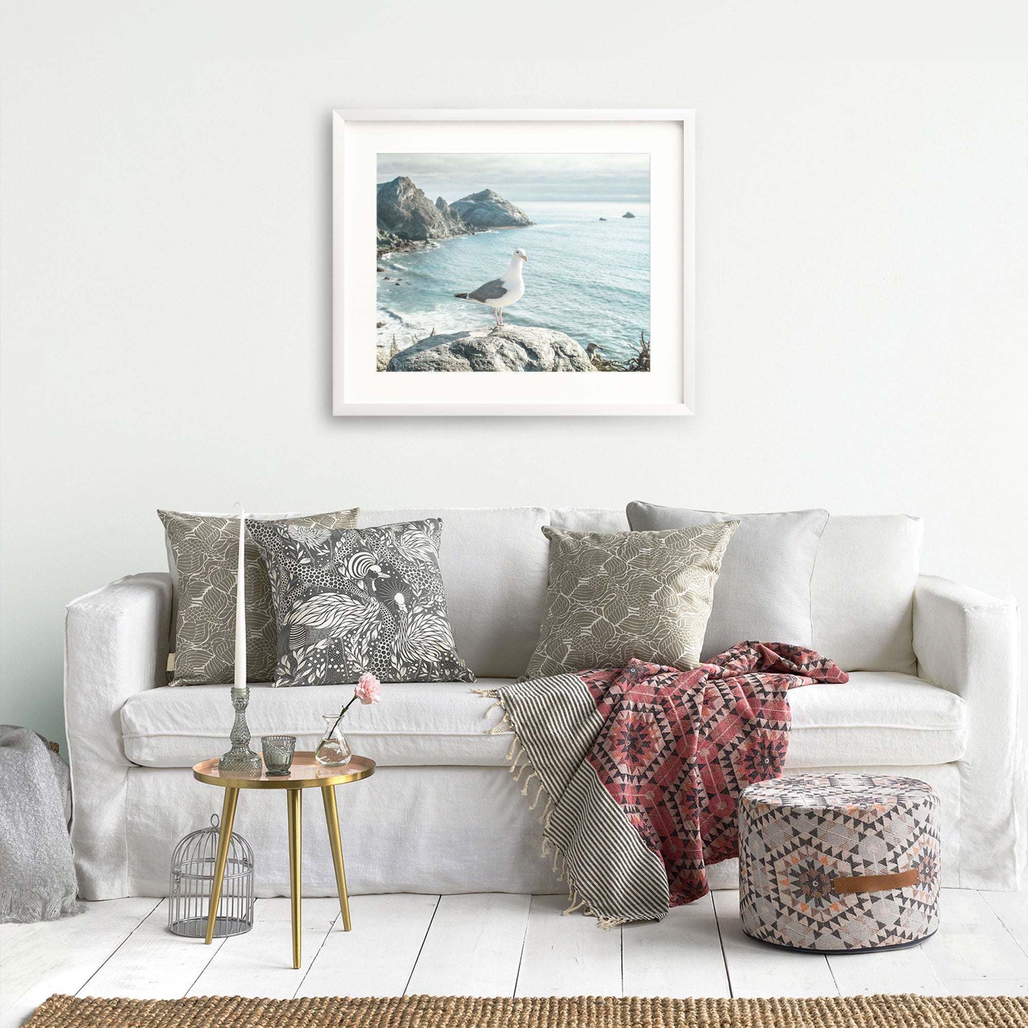A cozy living room with a white sofa adorned with patterned cushions, a red and gray throw blanket, a small round table with books and flowers, and a framed Offley Green Big Sur Landscape Print photograph on the wall.