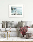A cozy living room with a white sofa adorned with patterned cushions, a red and gray throw blanket, a small round table with books and flowers, and a framed Offley Green Big Sur Landscape Print photograph on the wall.