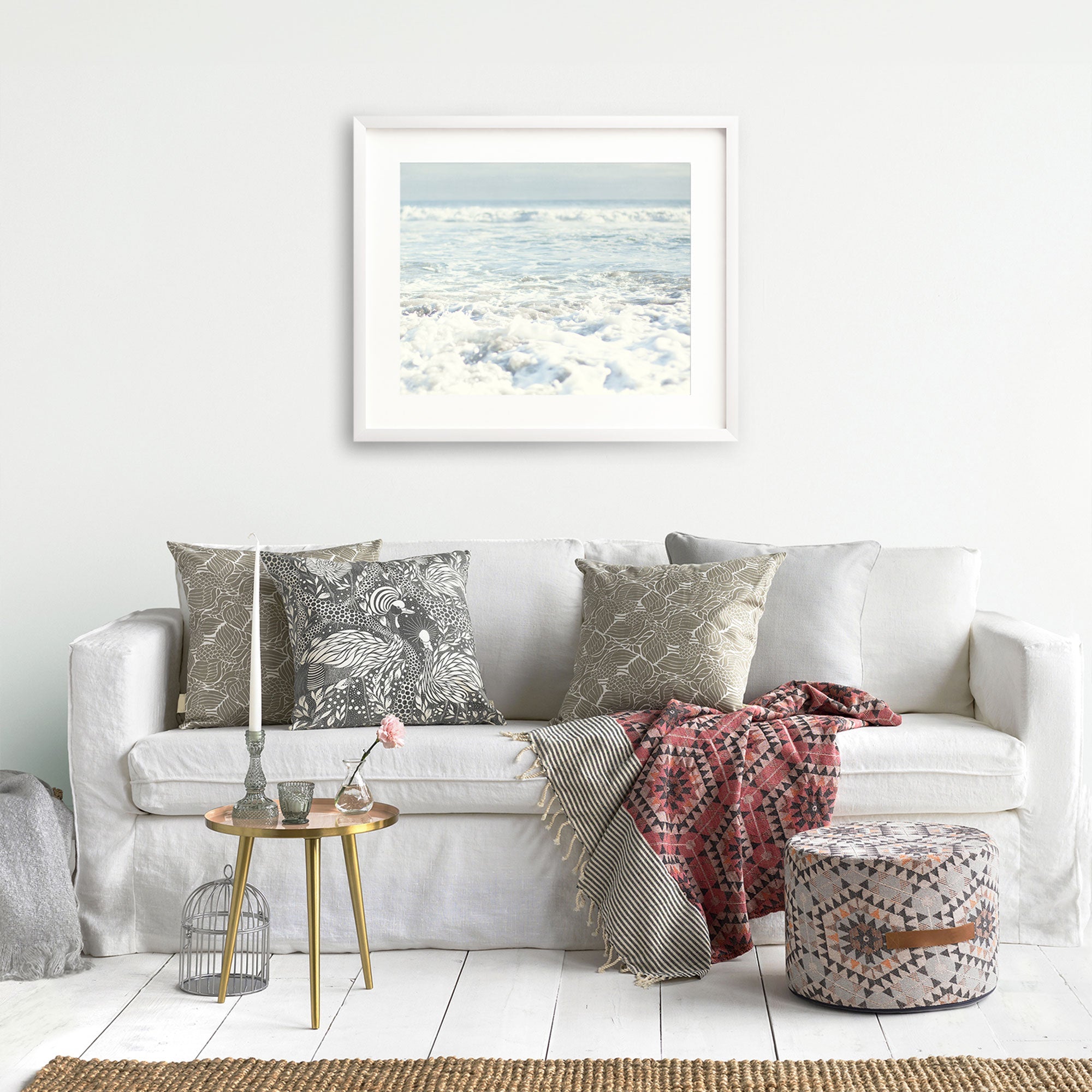 A stylish living room features a white couch with patterned pillows and a red throw. A round, gold side table holds a birdcage decor, candle, and glass vase with a rose. A patterned pouf sits on the floor, and an Offley Green Ocean Waves Print, 'Fresh Surf' printed on archival photographic paper hangs on a white wall.