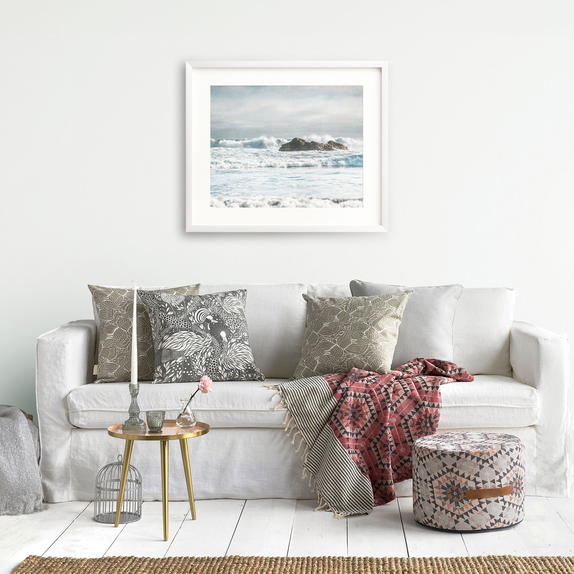 A cozy living room features a white sofa adorned with patterned throw pillows and a pink patterned blanket draped over the arm. A wooden side table with decorative items sits next to the sofa. On the wall hangs Offley Green's Seascape Coastal Art, 'Surf and Rocks', printed on archival photographic paper.