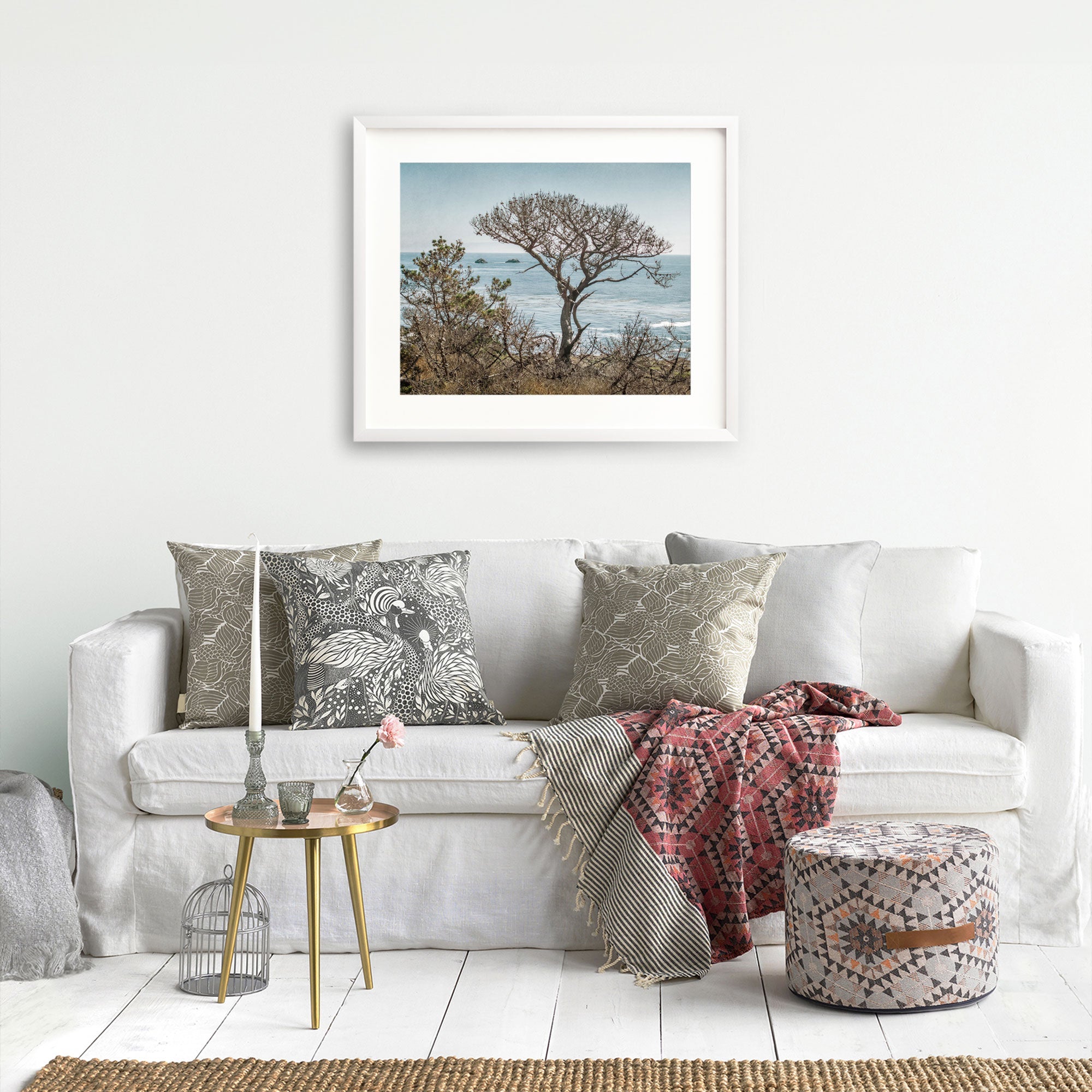 A cozy living room with a white sofa adorned with decorative pillows, a red patterned throw blanket, a small round table with a vase and books, and unframed Offley Green California Landscape Art in Big Sur, &#39;Wind Blown Tree&#39; photography on the wall.
