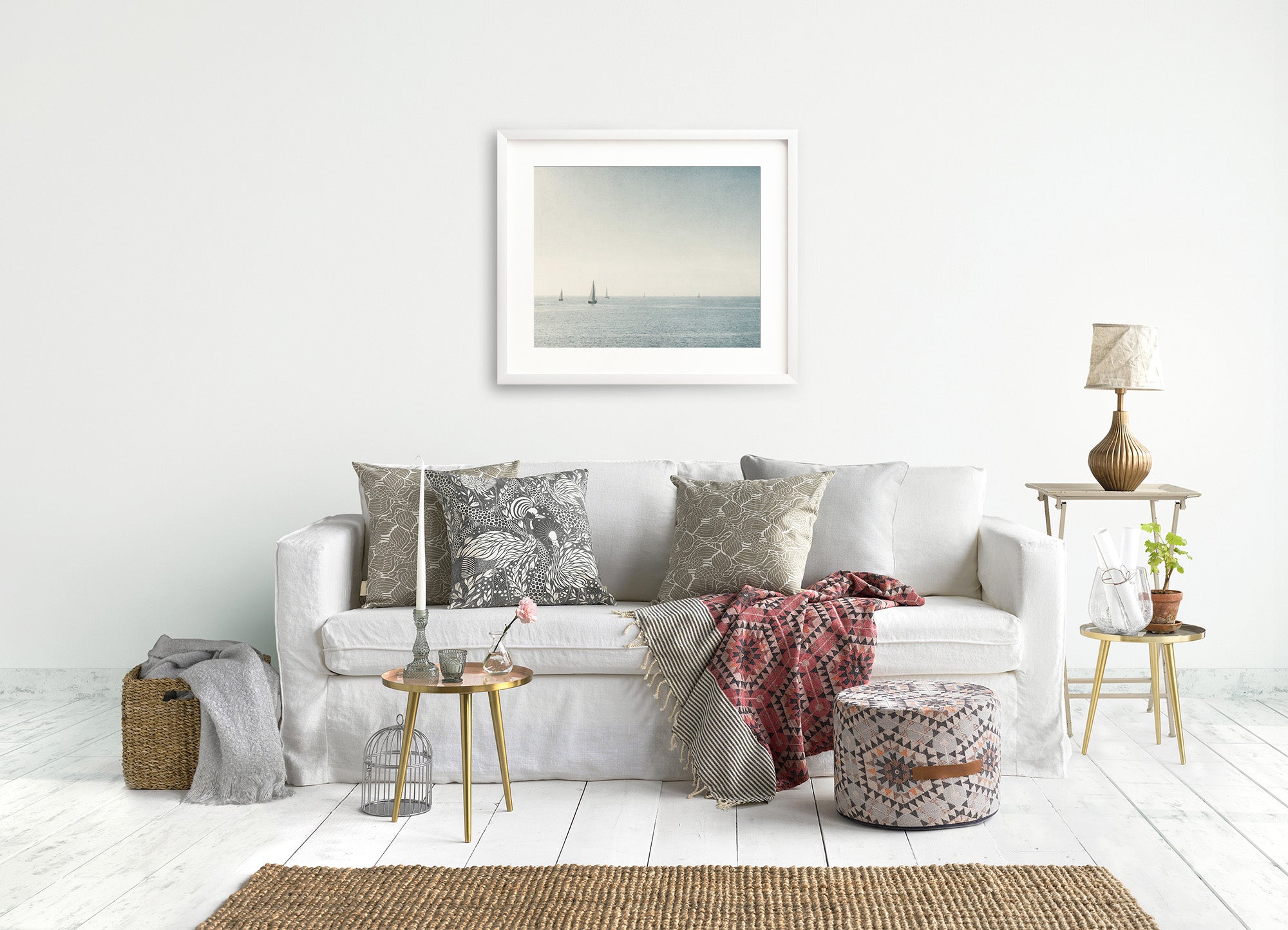 A cozy living room features a white sofa adorned with patterned cushions and throw blankets. An unframed *Moody Nautical Seascape Print, 'Sail Boats Approaching'* by Offley Green hangs above the sofa. Beside the sofa are small side tables with plants, a woven basket, a lamp, and decor items. The room has a light, airy ambiance.
