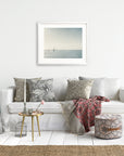 A cozy living room features a white sofa adorned with patterned cushions and throw blankets. An unframed *Moody Nautical Seascape Print, 'Sail Boats Approaching'* by Offley Green hangs above the sofa. Beside the sofa are small side tables with plants, a woven basket, a lamp, and decor items. The room has a light, airy ambiance.