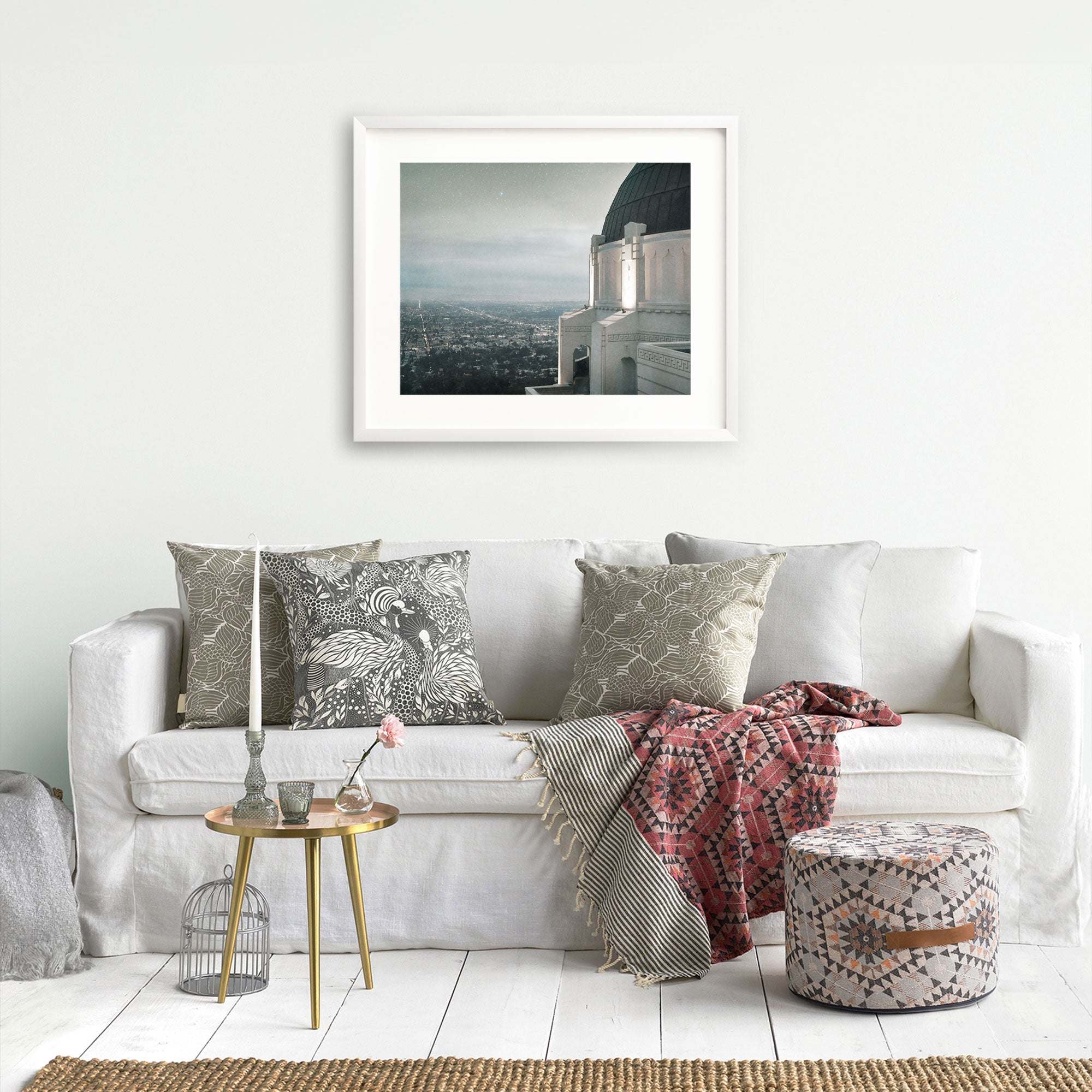 A minimalist living room featuring a white sofa adorned with patterned pillows, a small metallic side table with decor, a woven pouf, and an Offley Green archival photograph print of Griffith Observatory (&#39;The Sky At Night&#39;) hanging on the wall.