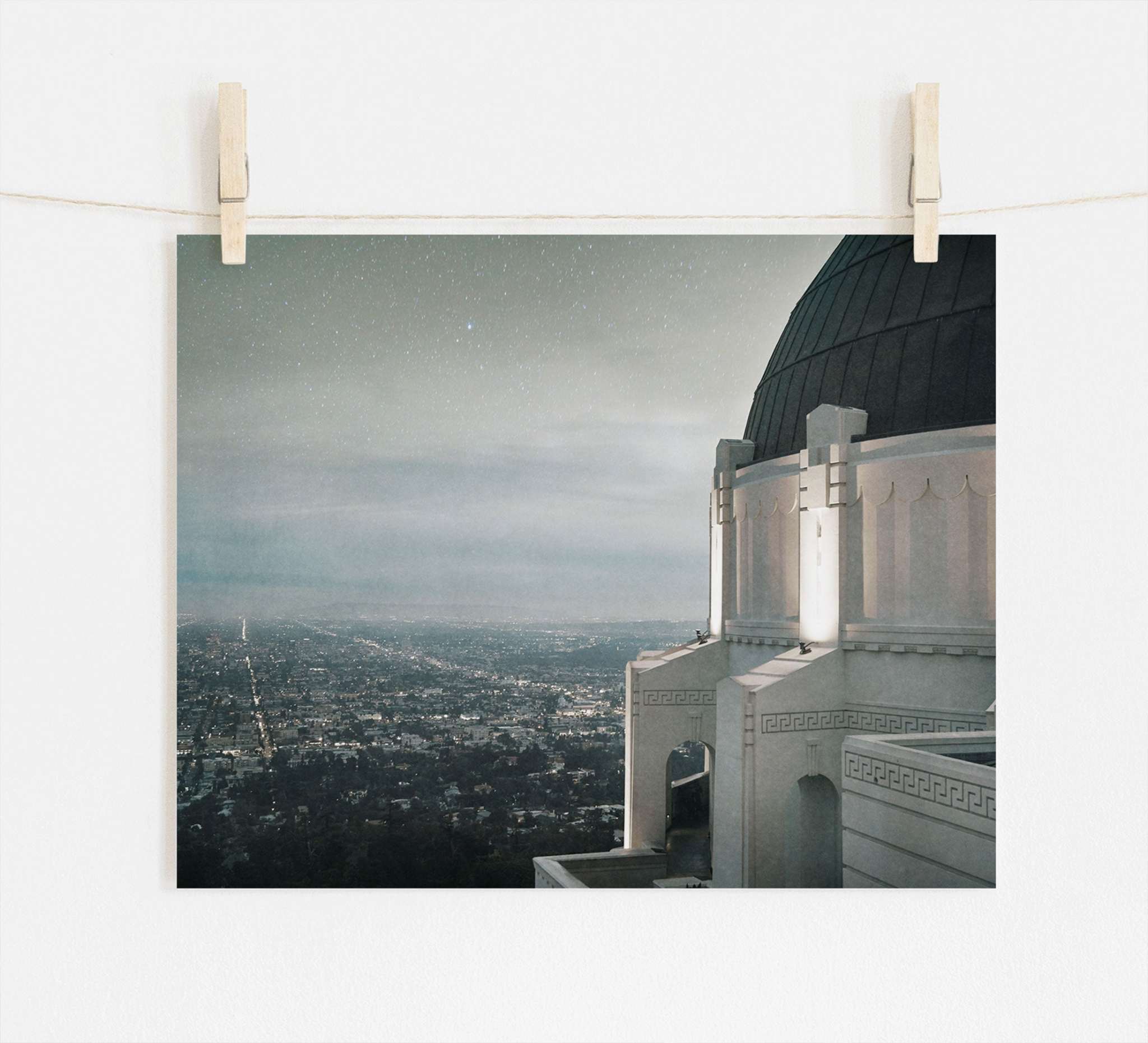 Photo of a cityscape at dusk pinned to a line, with Offley Green&#39;s Griffith Observatory Print, &#39;The Sky At Night&#39; in the foreground and twinkling city lights below a starry sky.