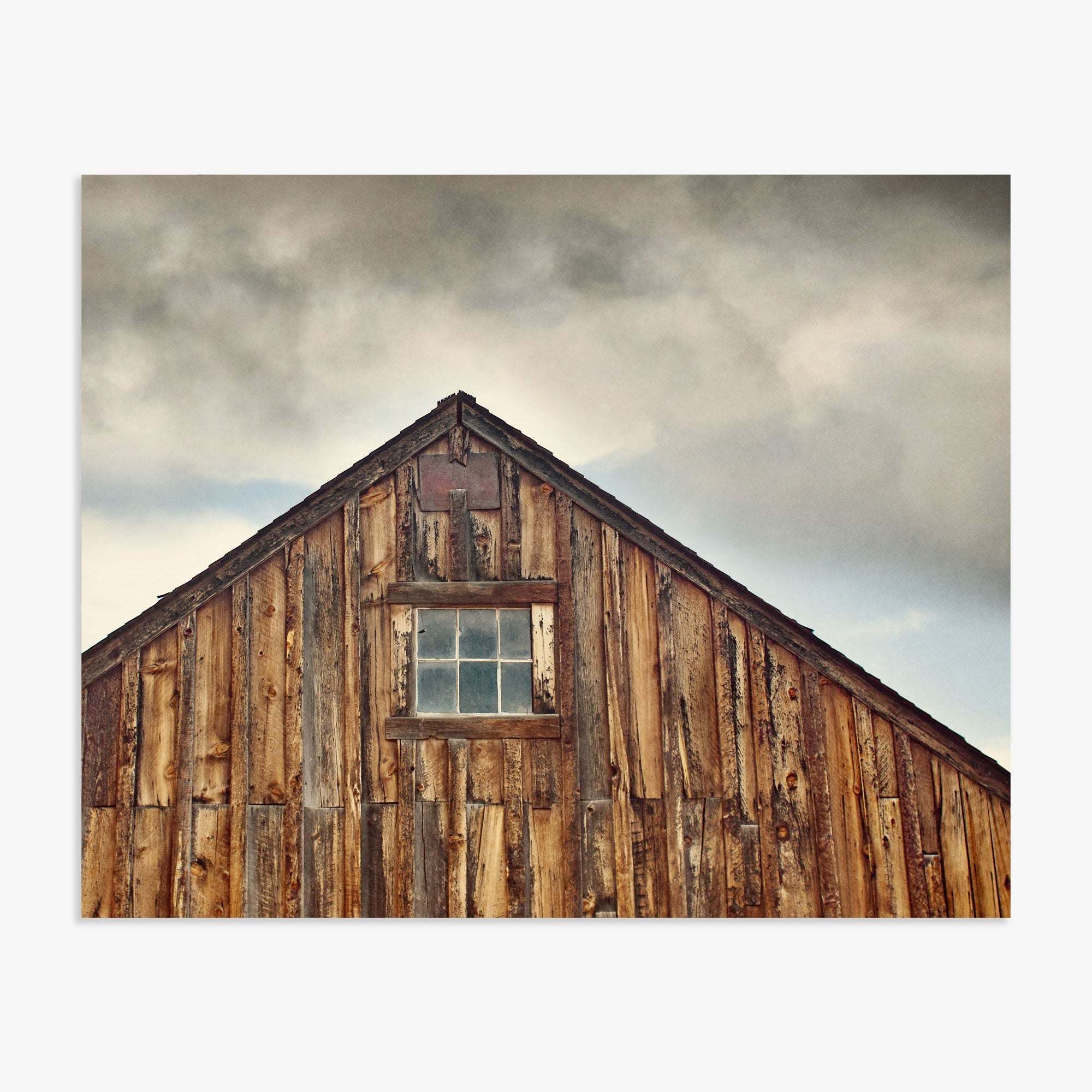 Offley Green&#39;s Farmhouse Rustic Print, &#39;Old Barn at Bodie&#39;, with weathered planks and a single window under a cloudy sky. The building&#39;s peak features intricate detailing against the backdrop of the Sierra Nevada mountains.