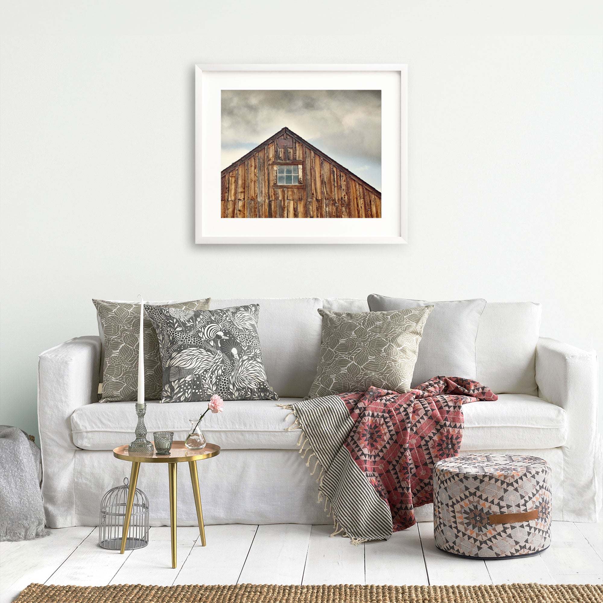 A cozy living room featuring a white sofa adorned with patterned throw pillows, a red and grey blanket, and a small round table with decorative items. Above the sofa, a framed photo of the Offley Green Farmhouse Rustic Print, &#39;Old Barn at Bodie&#39;.
