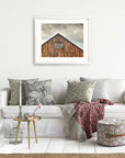 A cozy living room featuring a white sofa adorned with patterned throw pillows, a red and grey blanket, and a small round table with decorative items. Above the sofa, a framed photo of the Offley Green Farmhouse Rustic Print, 'Old Barn at Bodie'.