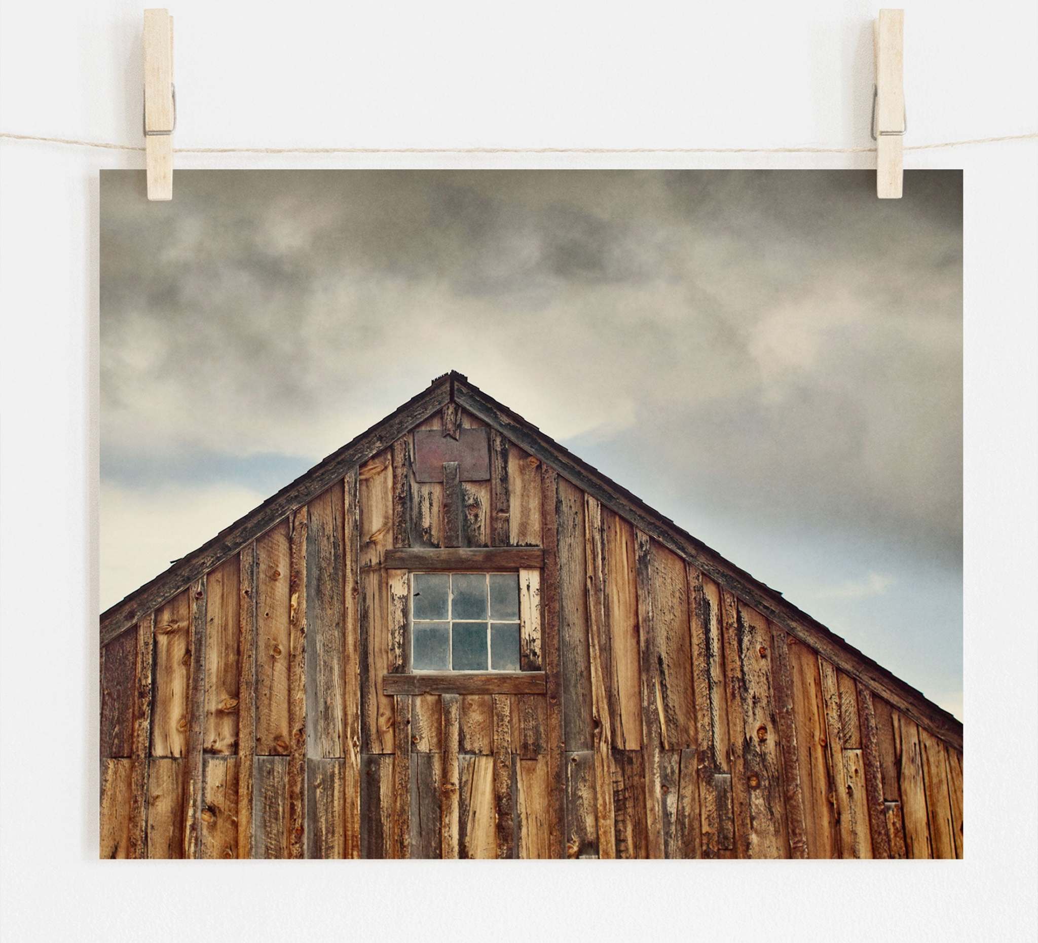 A photograph of a rustic farmhouse with a pitched roof and a single window, pinned up on white archival photographic paper by wooden clothespins. Dark clouds loom in the background. Offley Green&#39;s &#39;Old Barn at Bodie&#39; Farmhouse Rustic Print is a stunning addition to any decor.