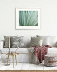 A cozy living room with a white sofa adorned with decorative pillows, a red patterned throw, a small round gold table with a vase and books, a pouf, and Offley Green's 'Strands and Spikes' Abstract Green Botanical Print.