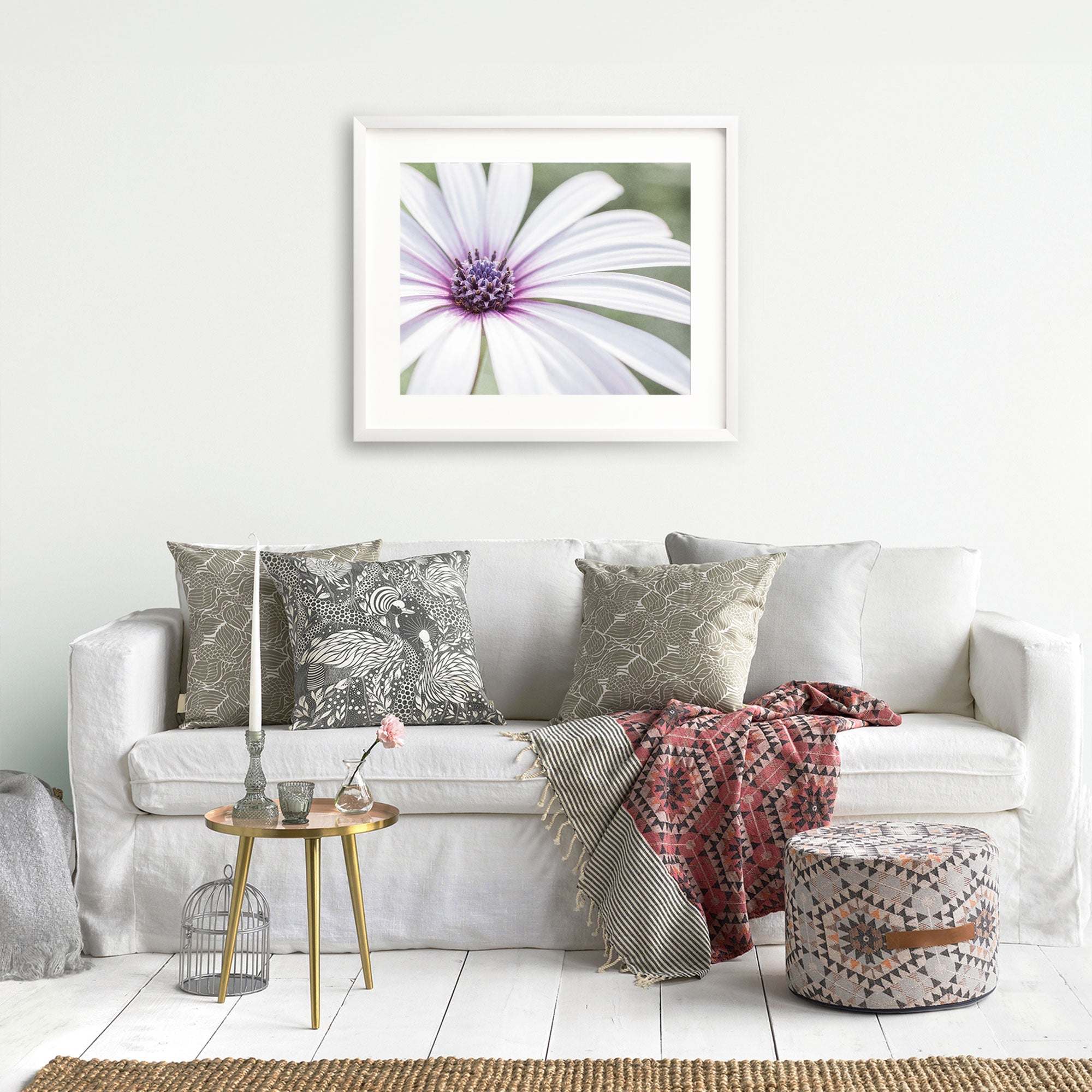 A cozy living room featuring a Large White Daisy Flower Print 'Bed of Petals' sofa adorned with various patterned cushions, a plush red blanket, a small round table with a vase and birdcage, an ottoman, and an unfr