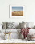 A cozy living room setup featuring a white sofa with decorative pillows, a glass side table, Offley Green California Central Coast Landscape Print 'Golden Ynez' on the wall, a red patterned throw blanket, and