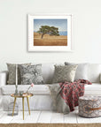 A cozy living room featuring a white sofa adorned with patterned cushions, a red throw blanket, a small round gold table with decorative items, and a framed California Oak Tree Print, 'Windswept' from Offley Green on the wall above.