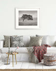 A cozy living room featuring a white sofa adorned with patterned pillows, a framed Offley Green Californian Oak Tree Landscape, 'Windswept (Black and White)' above it, a small round table with a vase, a birdcage.