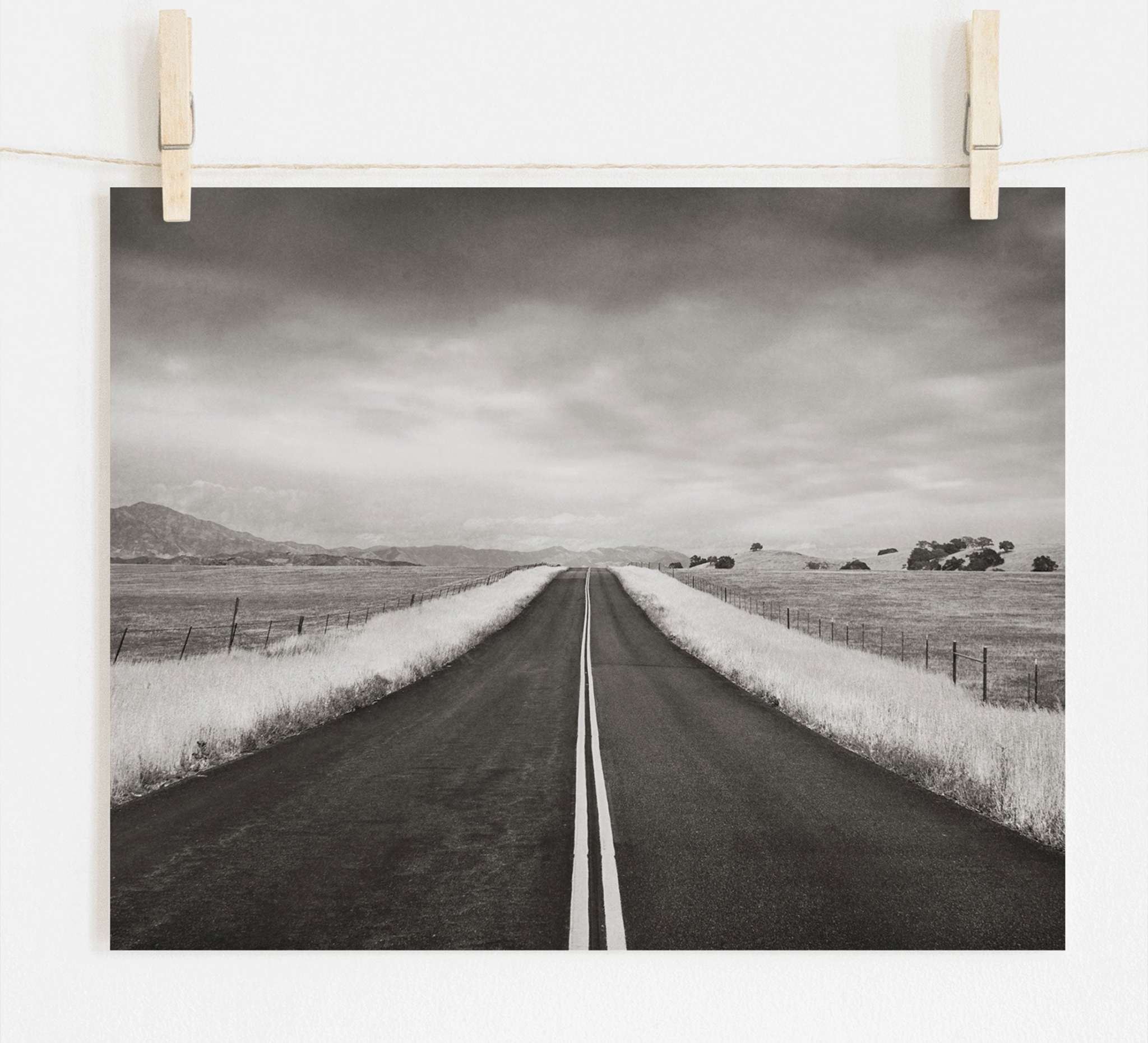 Offley Green's Black and White Rural Landscape Art, 'American Road Trip', printed on archival photographic paper, features an empty road extending into the distance under a cloudy sky, flanked by fields and mountains in sepia tones.