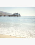 A tranquil coastal scene featuring Offley Green's 'Malibu Pier' California Beach Print extending into the ocean, with a large building at its end under a clear blue sky. Gentle waves touch a sandy beach in the foreground.