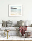 A modern living room featuring a white sofa adorned with patterned cushions, a cozy red blanket, and a small gold side table. On the wall above the sofa hangs a framed photo of California Beach Print by Offley Green.