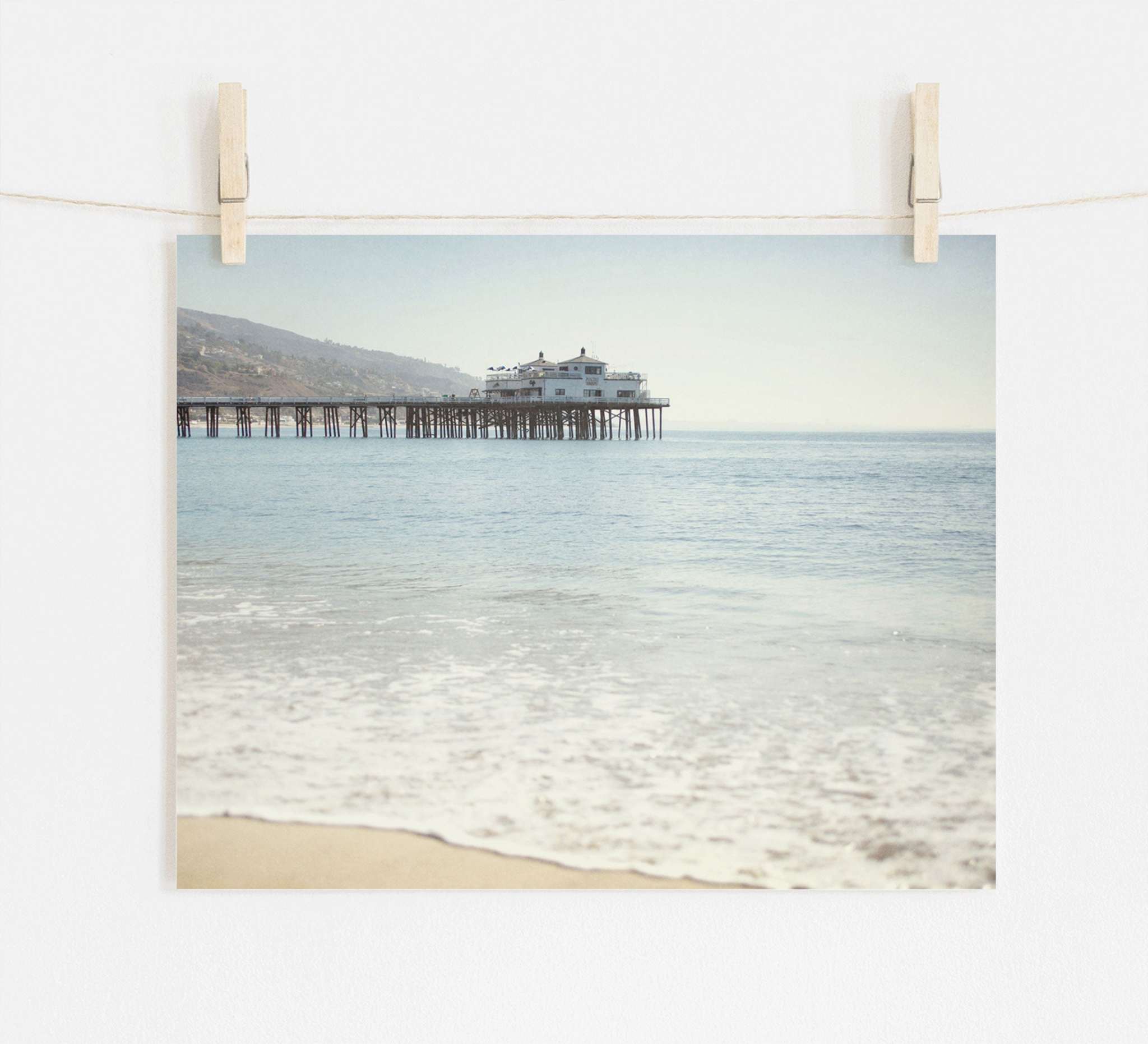 A photograph of Offley Green's 'Malibu Pier' California Beach Print extending into the ocean, pinned by wooden clothespins onto a string against a white wall. The calm sea and clear sky create a serene backdrop.