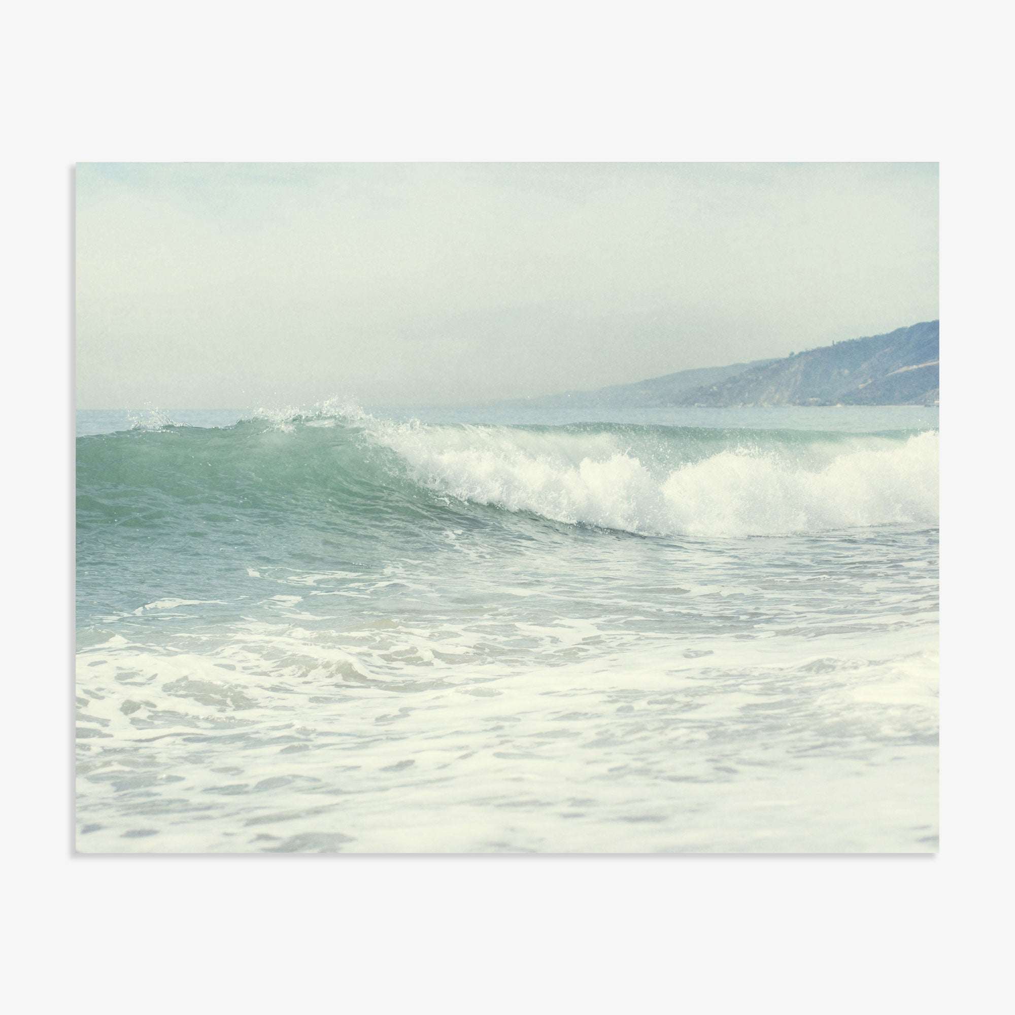 A serene Southern California beach scene depicting gentle waves rolling onto a sandy beach, with misty hills visible in the background under a soft, overcast sky. Offley Green's Coastal Print of a Breaking Wave 'Breaking Surf'.