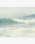 A serene Southern California beach scene depicting gentle waves rolling onto a sandy beach, with misty hills visible in the background under a soft, overcast sky. Offley Green's Coastal Print of a Breaking Wave 'Breaking Surf'.