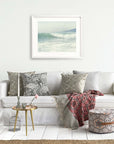 A cozy living room setup featuring a white sofa with patterned throw pillows, a draped red and gray blanket, a round gold side table with decor, a pouffe, and Offley Green's 'Breaking Surf' coastal print wall.
