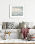 A cozy living room with a white sofa adorned with patterned cushions and a throw blanket exudes a serene atmosphere. A small round wooden table with decorative items sits in front of the sofa. On the white wall, a framed picture of Nautical Sail Boat Art, 'Sailing Into Rain' by Offley Green adds an elegant touch. A patterned pouf sits nearby.