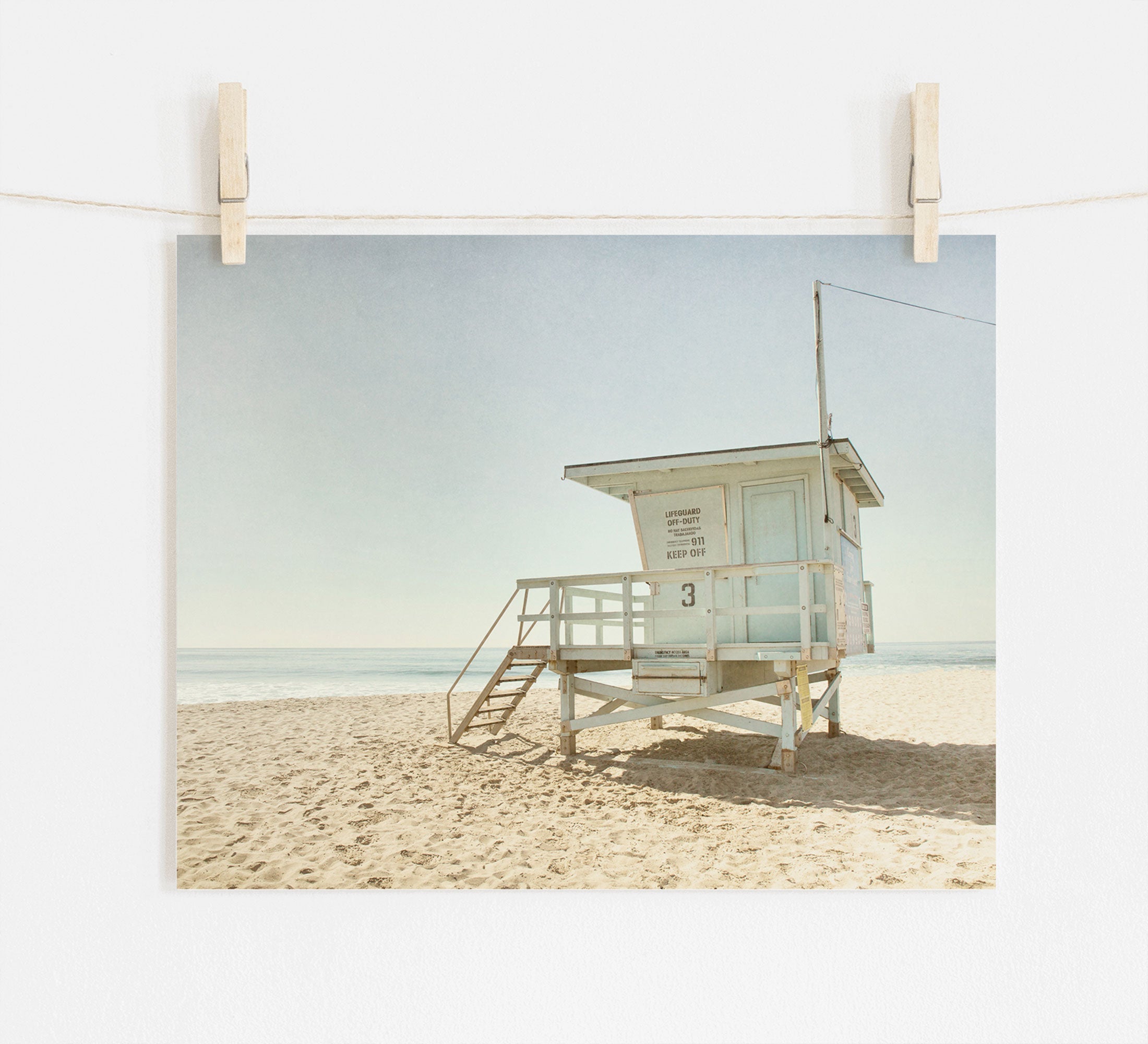A photo of Offley Green&#39;s California Summer Beach Art, &#39;Malibu Lifeguard Tower&#39;, numbered &quot;3&quot; on Malibu beach, pinned by wooden clothespins on a line, set against a scene with soft sand and a clear sky.