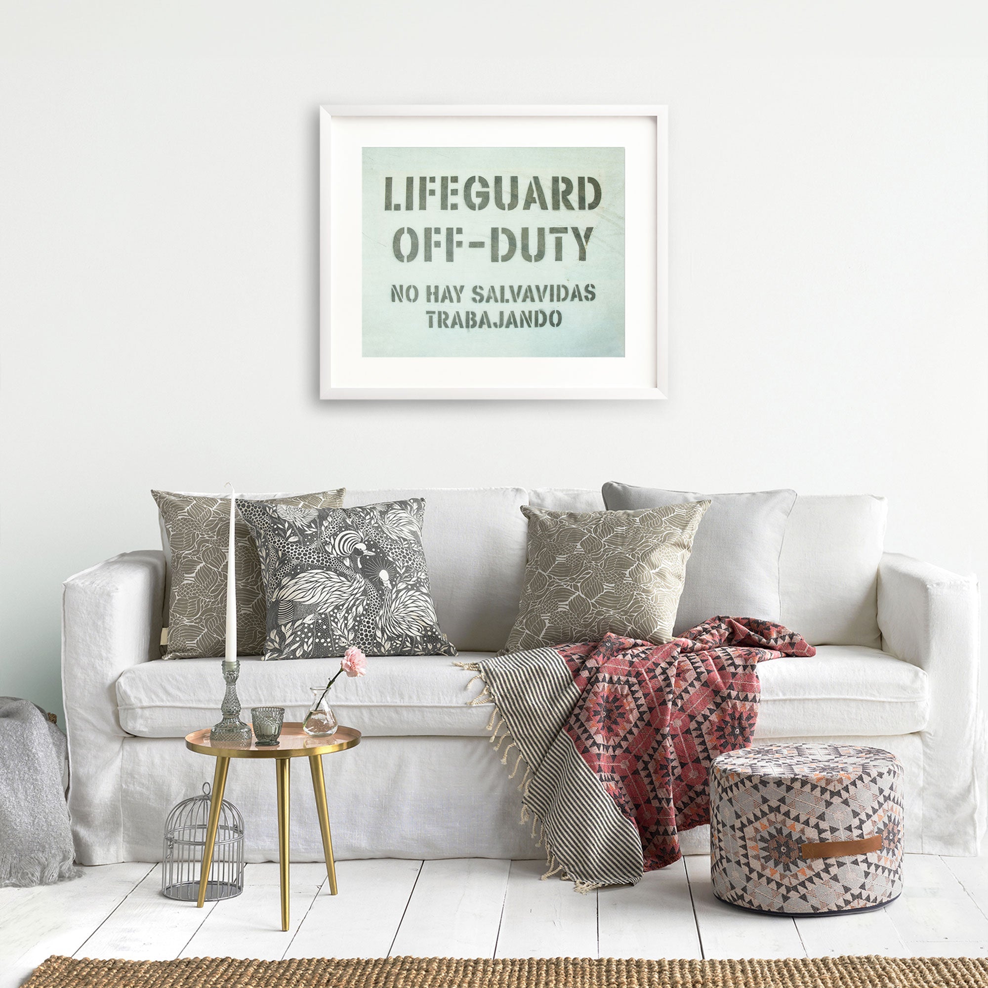 A cozy living room with a white sofa adorned with various patterned cushions. Above the sofa hangs Offley Green's Teal Blue Surfer Beach Print, 'Off Duty'. A small round side table with a candle and birdcage decor is placed nearby.