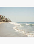 A serene Malibu coastline scene with gentle waves lapping at the shore and a row of multistory beach houses on stilts, lining the sandy coast under a clear sky featuring the Malibu Beach House Print by Offley Green.